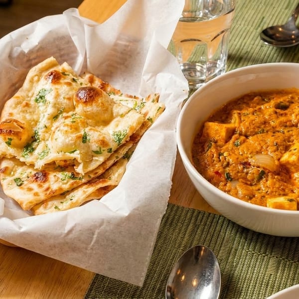 A basket of naan bread and a bowl of paneer curry on a table with a glass of water.