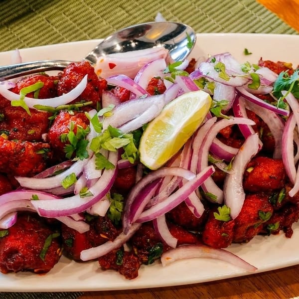 Bright red chicken pakora with sliced red onions, cilantro, and a lemon wedge on a white plate.
