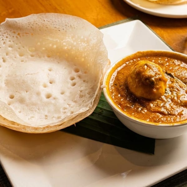 A bowl of egg curry and two appam pancakes on a white plate with a green placemat.